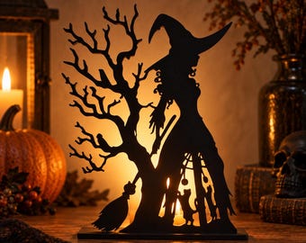 Puede incluir: Una decoración de silueta de metal negro que muestra a una bruja con sombrero puntiagudo de pie junto a un árbol desnudo, con una escoba y dos gatos en la base. El diseño recortado permite que la luz cálida pase a través de las formas.