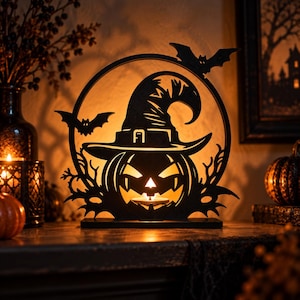 May include: A black circular decorative cutout featuring a carved pumpkin wearing a witch hat, accented with two bats and branch-like silhouettes. The design is illuminated from behind, casting a warm glow through the cutouts of the face.