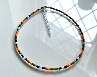 May include: A beaded necklace featuring a repeating pattern of small, round, multi-colored glass beads in red, orange, yellow, green, blue, white, black, and metallic gold. The necklace includes a silver-toned metal lobster clasp and chain.