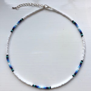 Puede incluir: Un collar gargantilla de cuentas blancas con detalles azules y negros. El collar tiene un cierre plateado.