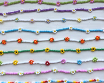 Op de afbeelding: Negen kralenarmbanden met daisy bloemcharms in verschillende kleuren, waaronder groen, paars, wit, lichtblauw, oranje en roze.