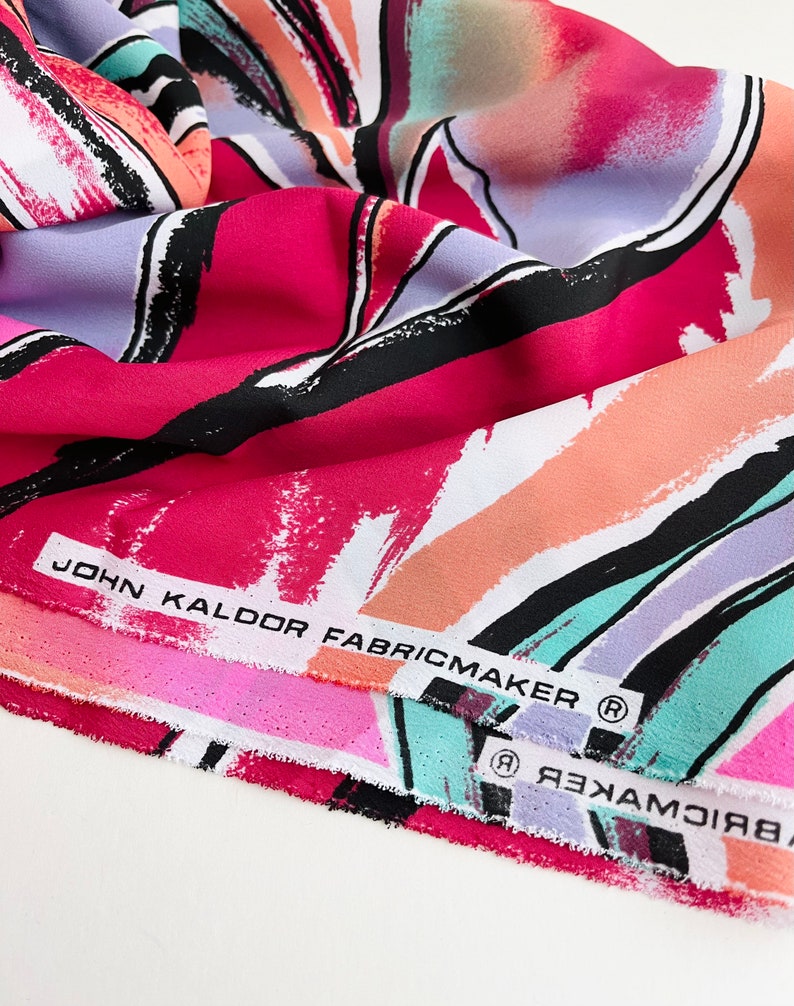 May include: Close-up of a folded fabric with an abstract print in shades of pink, orange, blue, and green. The fabric has a black and white brushstroke design. The label reads "John Kaldor Fabricmaker (R)".