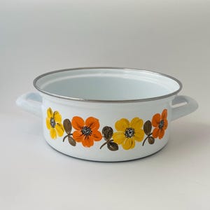 May include: White enamelware pot with two handles and a silver rim. The pot is decorated with a repeating pattern of orange and yellow flowers with brown leaves. The interior of the pot is light blue.