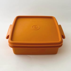 May include: An orange square Tupperware container with a matching lid. The lid has a circular indentation in the center and two handles on the sides. The container is made of durable plastic.