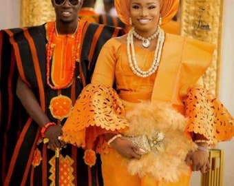 May include: A couple in traditional Nigerian wedding attire. The woman is wearing a bright orange dress with intricate lace detailing and a matching head wrap. The man is wearing a black and orange striped robe with a matching cap.