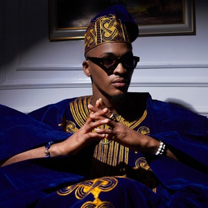 May include: A man wearing a blue velvet robe with gold embroidery and a blue hat with gold embroidery. He is wearing sunglasses and has his hands clasped in his lap.