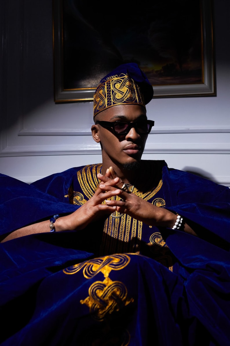 May include: A man wearing a blue velvet robe with gold embroidery and a blue hat with gold embroidery. He is wearing sunglasses and has his hands clasped in his lap.