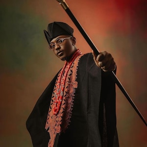 May include: A man wearing a black outfit with intricate embroidery and a red beaded necklace holds a silver-tipped walking stick. He is wearing a black hat and glasses.