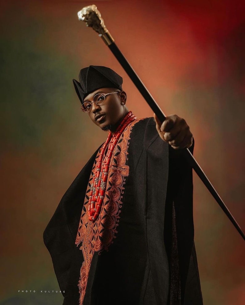 May include: A man wearing a black outfit with intricate embroidery and a red beaded necklace holds a silver-tipped walking stick. He is wearing a black hat and glasses.