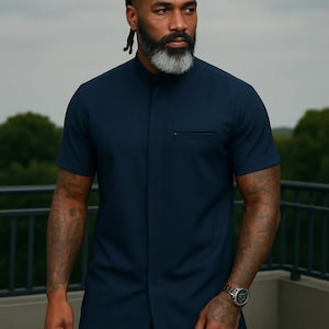 May include: A man wearing a dark blue short-sleeved collared shirt with a single button closure. He has a gray beard and braided hair.