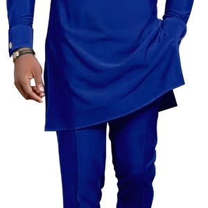 May include: A royal blue two-piece suit with a long-sleeved tunic and matching trousers. The tunic features a white panel with three black stripes and a small crown emblem. The suit is a modern take on traditional African attire.