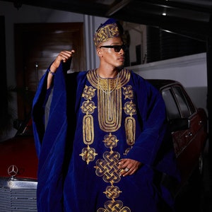 May include: A man wearing a blue velvet robe with gold embroidery. The robe has a high neckline and long sleeves. The man is wearing a gold hat and sunglasses.