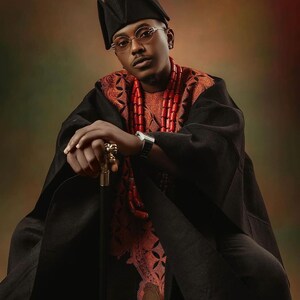 May include: A man wearing a black robe with a red beaded necklace and a black hat. He is holding a gold-tipped walking stick and looking directly at the camera.