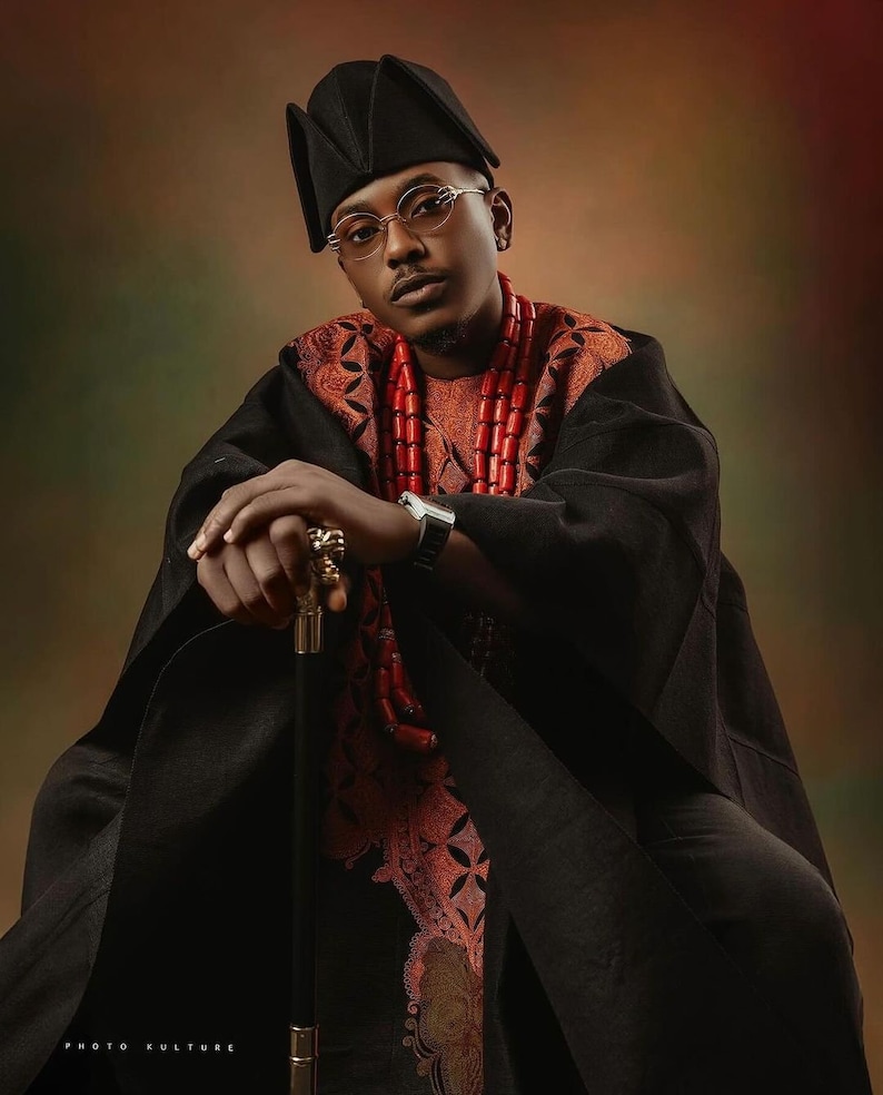 May include: A man wearing a black robe with a red beaded necklace and a black hat. He is holding a gold-tipped walking stick and looking directly at the camera.