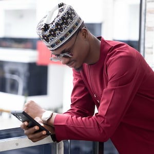 May include: A man wearing a red shirt and a patterned cap is looking down at a black smartphone. He is standing on a balcony with a metal railing.