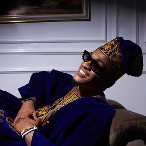 May include: A man wearing a blue velvet robe with gold embroidery, a black hat with gold trim, and black sunglasses. He is sitting on a couch and smiling.