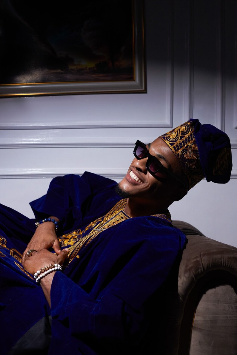 May include: A man wearing a blue velvet robe with gold embroidery, a black hat with gold trim, and black sunglasses. He is sitting on a couch and smiling.