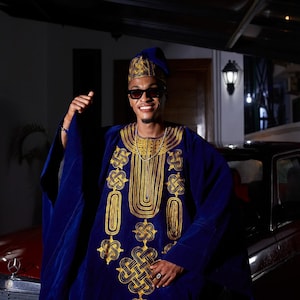 May include: A man wearing a blue and gold patterned robe, a blue hat, and sunglasses. He is standing in front of a car and smiling.