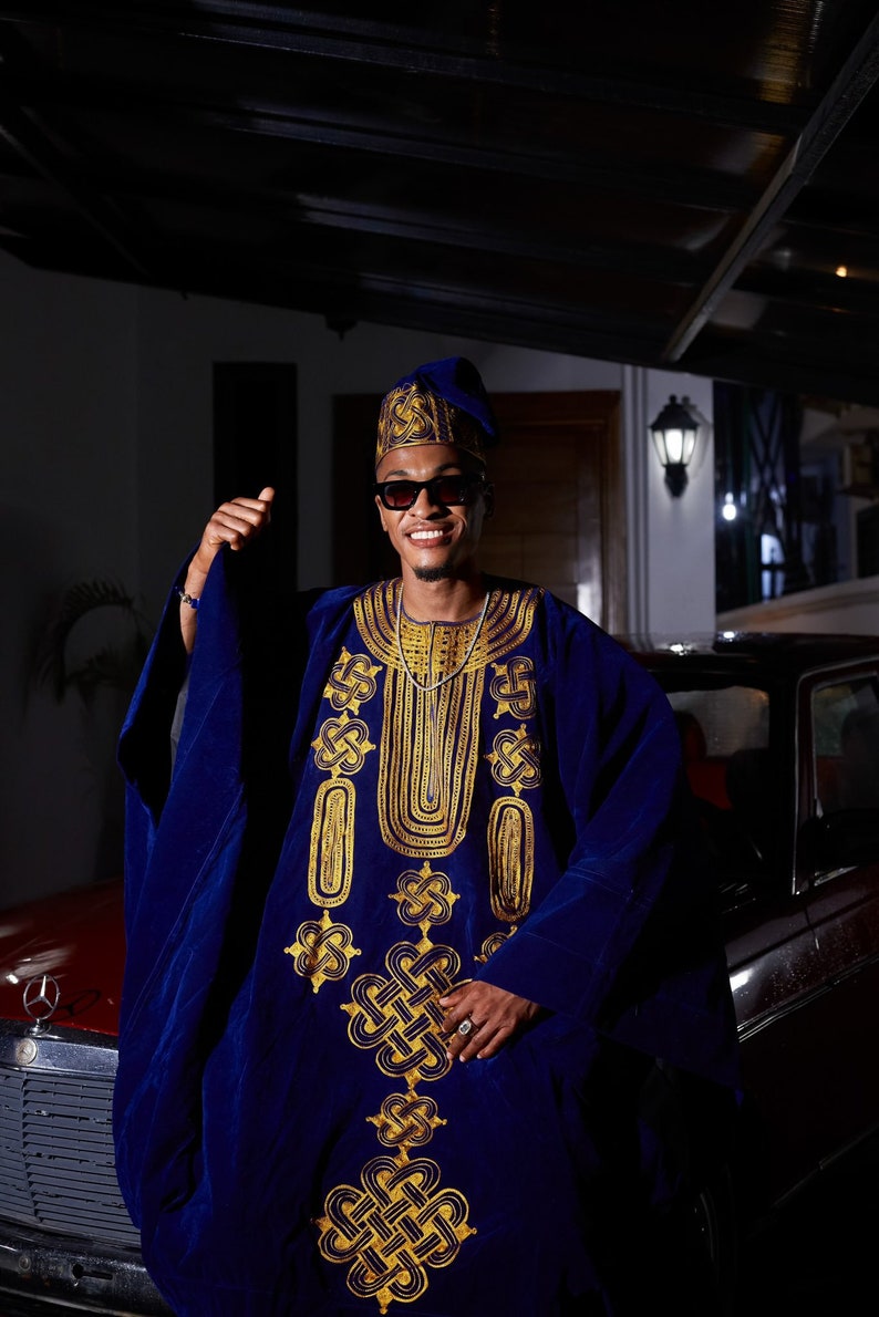 May include: A man wearing a blue and gold patterned robe, a blue hat, and sunglasses. He is standing in front of a car and smiling.