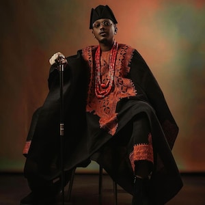 May include: A man wearing a black robe with intricate orange embroidery and a red beaded necklace. He is sitting on a chair and holding a black walking stick. The background is a gradient of orange and green.