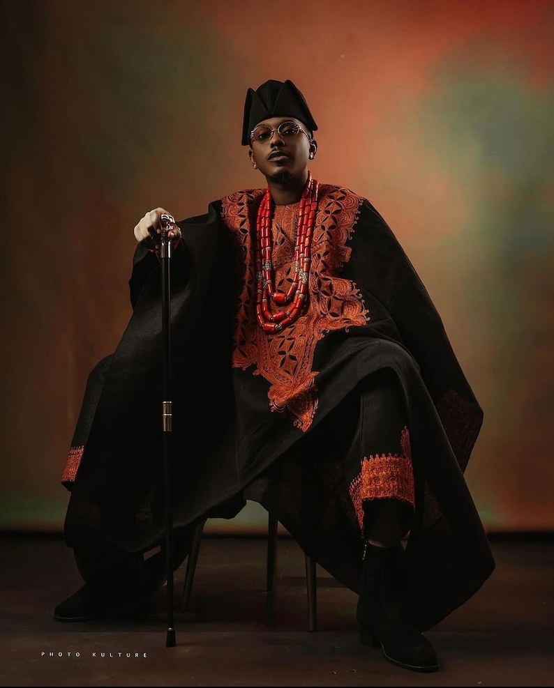 May include: A man wearing a black robe with intricate orange embroidery and a red beaded necklace. He is sitting on a chair and holding a black walking stick. The background is a gradient of orange and green.