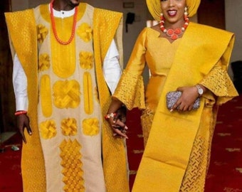 May include: A couple wearing traditional Nigerian wedding attire. The woman is wearing a yellow dress with a matching head wrap and jewelry. The man is wearing a yellow and beige embroidered robe with a red beaded necklace.