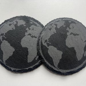 May include: Two round, dark gray slate coasters. Each coaster features a detailed, etched map of the world. The coasters are designed with a natural, textured edge and are placed side by side on a white surface.