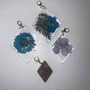 May include: Four clear resin charms in a diamond shape, each with a different dried flower or design. The charms have gold or silver-colored clasps. The flowers are blue, purple, and brown.