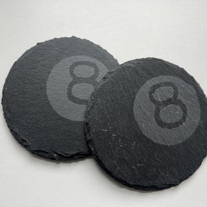 May include: Two round, black slate coasters. Each coaster features a gray, etched design of the number eight, resembling a billiard ball. The coasters have a rough, natural edge and are set against a white background.