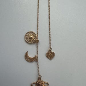 May include: A gold-colored necklace featuring celestial charms. The necklace includes a sun, crescent moon, planet, and heart pendant. The sun and moon have intricate cut-out designs, while the planet has a star. The heart has a heart-shaped design.