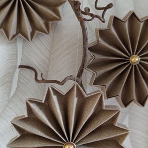 May include: Decorative paper starburst ornaments in shades of brown and tan, each with a gold-colored center. The starbursts are attached to a brown, twig-like branch, creating a natural, rustic aesthetic. The background is a textured, off-white surface.