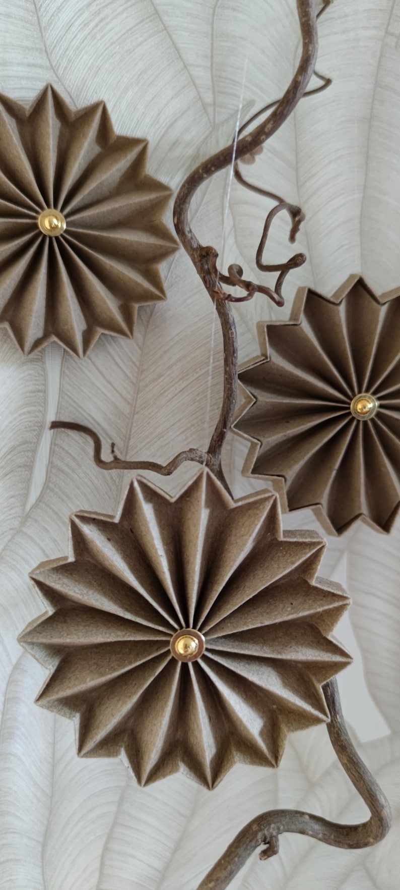 May include: Decorative paper starburst ornaments in shades of brown and tan, each with a gold-colored center. The starbursts are attached to a brown, twig-like branch, creating a natural, rustic aesthetic. The background is a textured, off-white surface.
