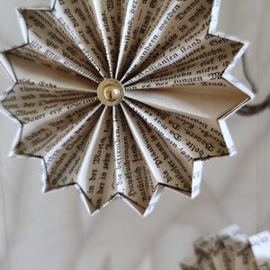May include: Handmade paper star ornaments, crafted from vintage book pages. Each star features a gold-colored button center and is suspended by a thin string. The paper has a beige hue with black text, creating a rustic, literary aesthetic.