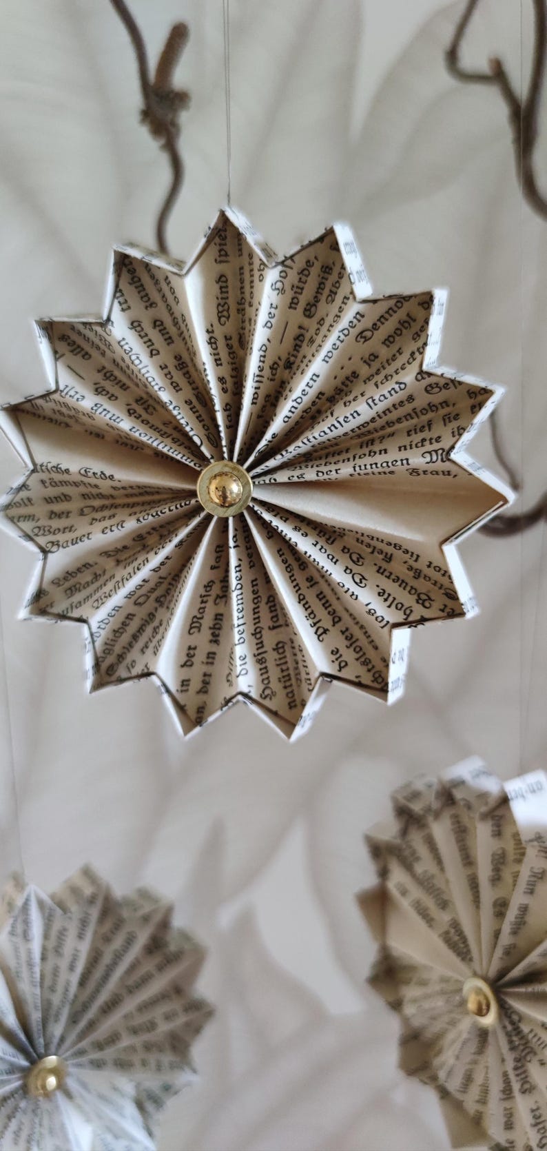 May include: Handmade paper star ornaments, crafted from vintage book pages. Each star features a gold-colored button center and is suspended by a thin string. The paper has a beige hue with black text, creating a rustic, literary aesthetic.