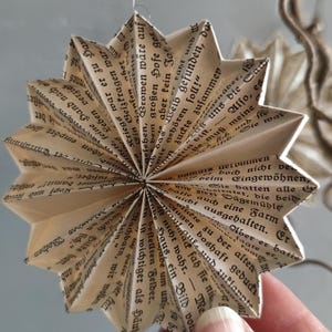 May include: A handmade star ornament crafted from vintage book pages. The paper star, with its intricate folds and visible text, hangs from a thin string, showcasing a unique, eco-friendly design. The neutral tones and literary theme create a rustic, artistic aesthetic.