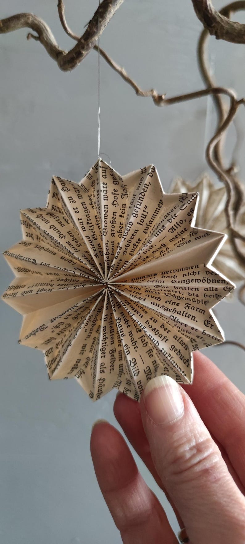 May include: A handmade star ornament crafted from vintage book pages. The paper star, with its intricate folds and visible text, hangs from a thin string, showcasing a unique, eco-friendly design. The neutral tones and literary theme create a rustic, artistic aesthetic.