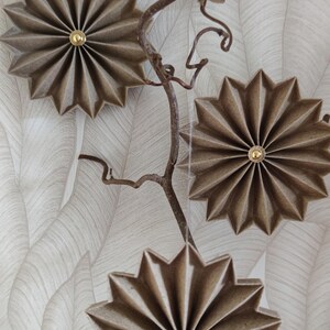 May include: Three decorative, star-shaped wall hangings with a gold center, made of textured, brown paper. They are attached to a thin, brown branch against a white and gray leaf-patterned background.