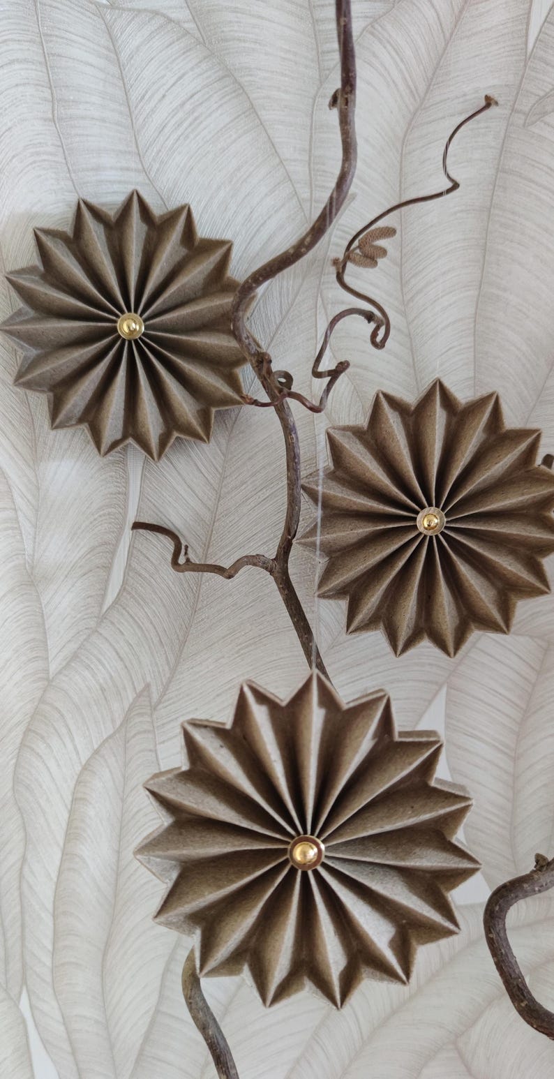 May include: Three decorative, star-shaped wall hangings with a gold center, made of textured, brown paper. They are attached to a thin, brown branch against a white and gray leaf-patterned background.