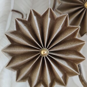 May include: Close-up of handmade, star-shaped ornaments. Each ornament is crafted from folded, light brown paper and features a small, gold-colored center. The ornaments are suspended from a natural-looking branch, suggesting a festive or decorative theme.