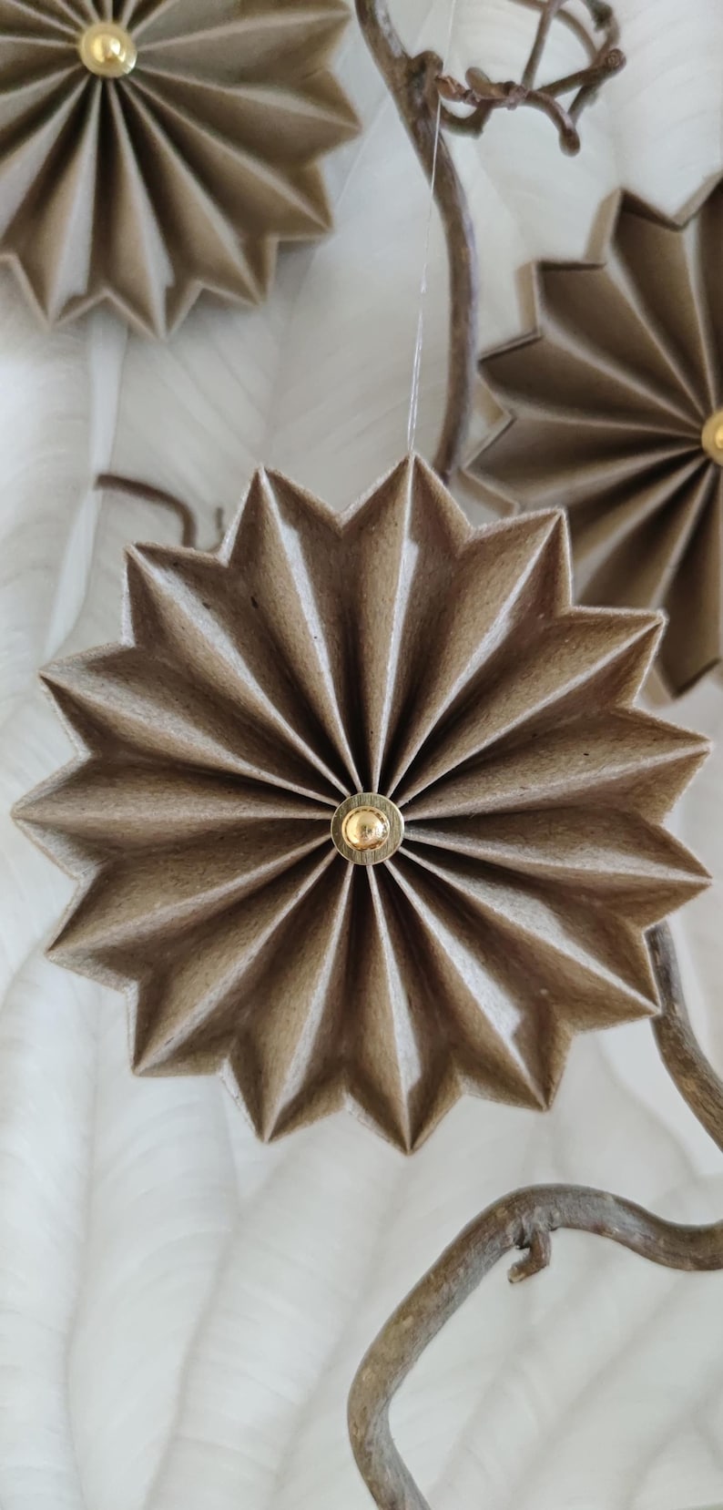 May include: Close-up of handmade, star-shaped ornaments. Each ornament is crafted from folded, light brown paper and features a small, gold-colored center. The ornaments are suspended from a natural-looking branch, suggesting a festive or decorative theme.