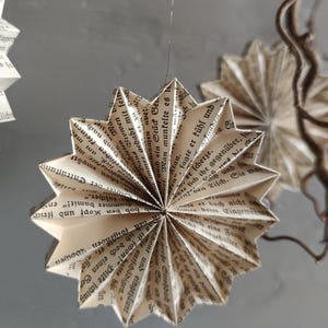 May include: Handmade paper star ornaments, crafted from vintage book pages. These unique decorations feature a starburst design, suspended by a thin string, and add a touch of rustic charm to any space.