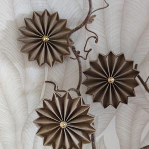 May include: Three brown, star-shaped paper ornaments with gold centers hang from a brown branch against a white and gray leaf-patterned background. The ornaments have a pleated design.