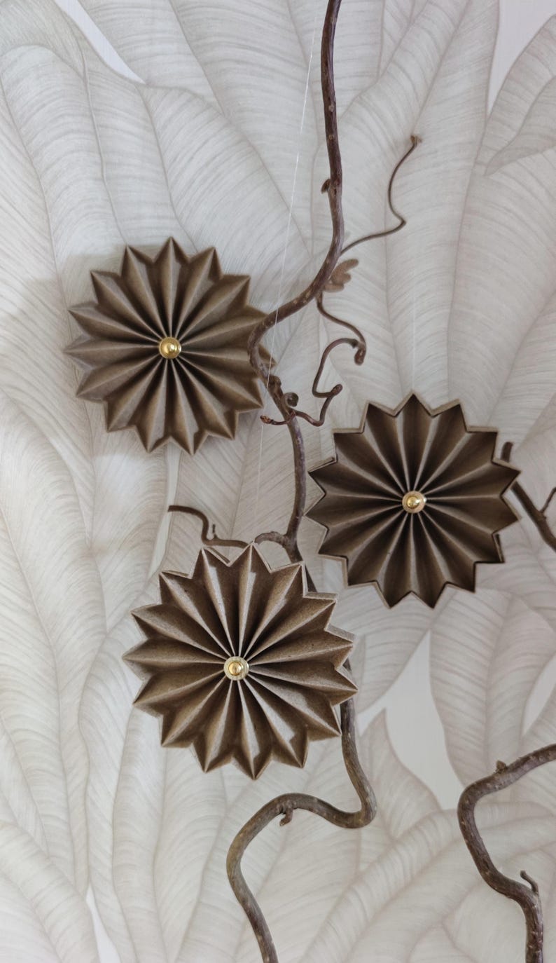 May include: Three brown, star-shaped paper ornaments with gold centers hang from a brown branch against a white and gray leaf-patterned background. The ornaments have a pleated design.