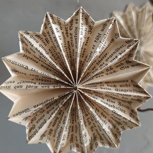 May include: A handmade, star-shaped ornament crafted from the pages of a vintage book. The paper is folded into a multi-pointed design, with visible text adding a unique, rustic charm. The ornament hangs from a thin string.