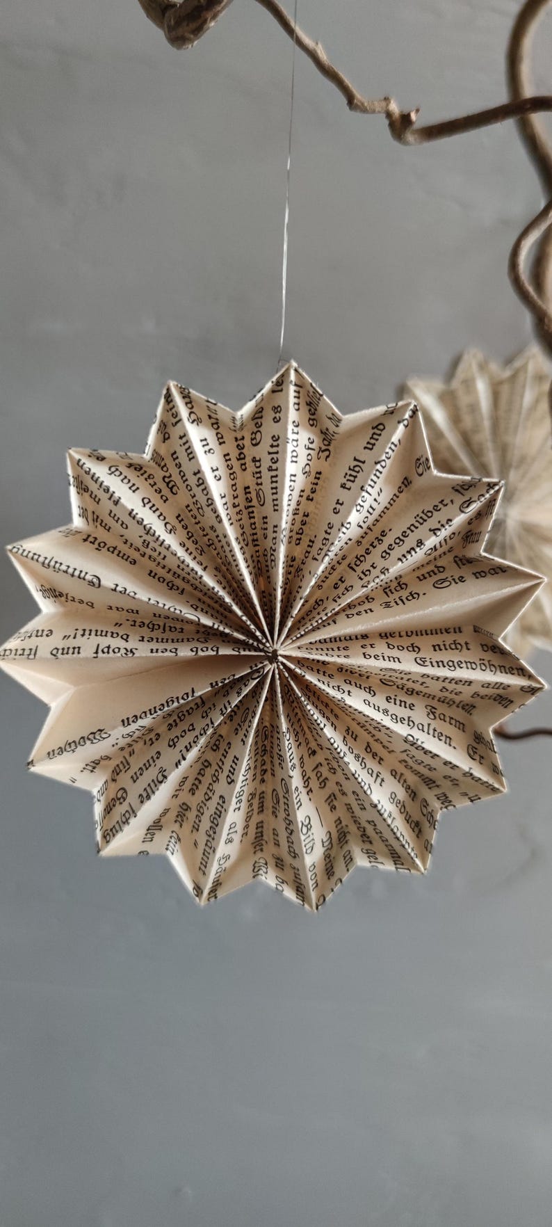 May include: A handmade, star-shaped ornament crafted from the pages of a vintage book. The paper is folded into a multi-pointed design, with visible text adding a unique, rustic charm. The ornament hangs from a thin string.