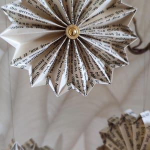 May include: Paper star ornaments made from vintage book pages. Each star is folded and has a gold-colored button in the center. The stars are hanging against a neutral background, creating a rustic, literary aesthetic.