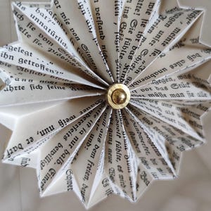 May include: A handmade paper star ornament, crafted from pages of a book. The star has a gold center and is suspended by a thin string. The paper is aged, with visible text, creating a vintage aesthetic.