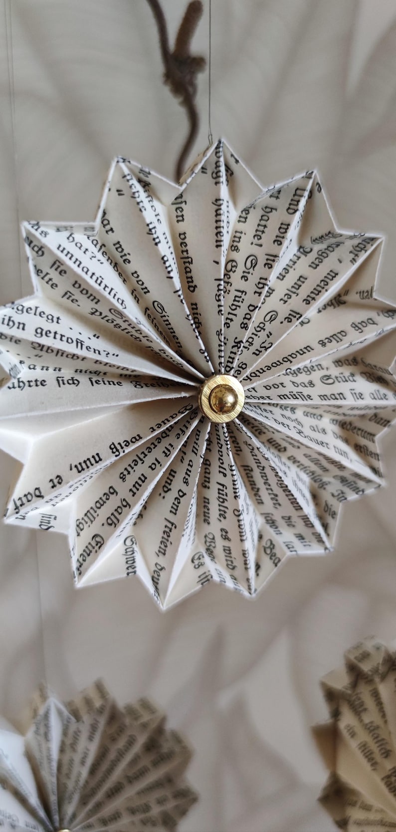 May include: A handmade paper star ornament, crafted from pages of a book. The star has a gold center and is suspended by a thin string. The paper is aged, with visible text, creating a vintage aesthetic.