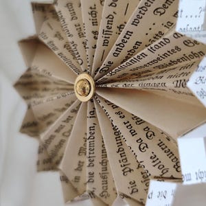 May include: A handmade paper ornament shaped like a starburst, crafted from vintage book pages. The pages display German text and are held together with a gold-colored button. A unique, eco-friendly decoration.