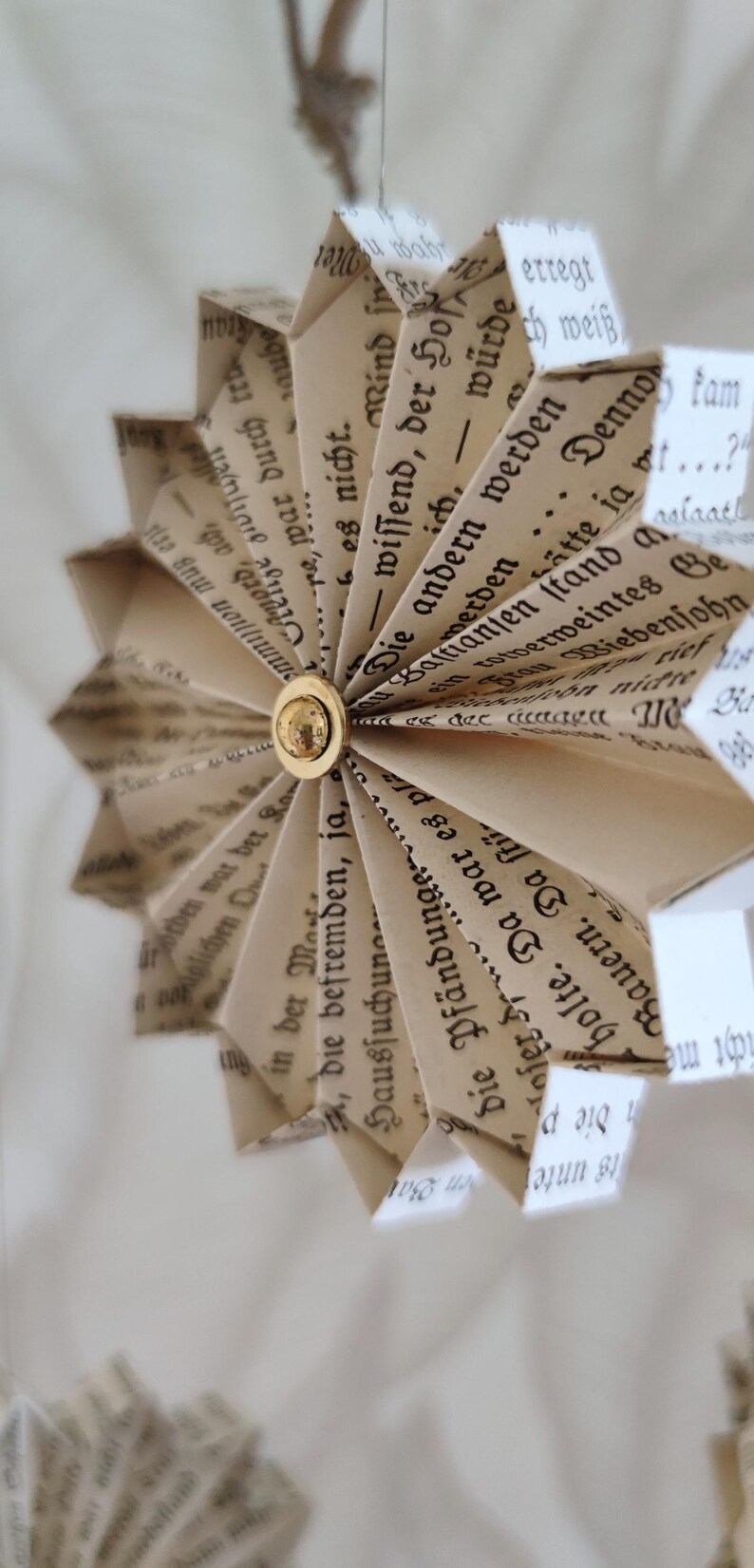 May include: A handmade paper ornament shaped like a starburst, crafted from vintage book pages. The pages display German text and are held together with a gold-colored button. A unique, eco-friendly decoration.
